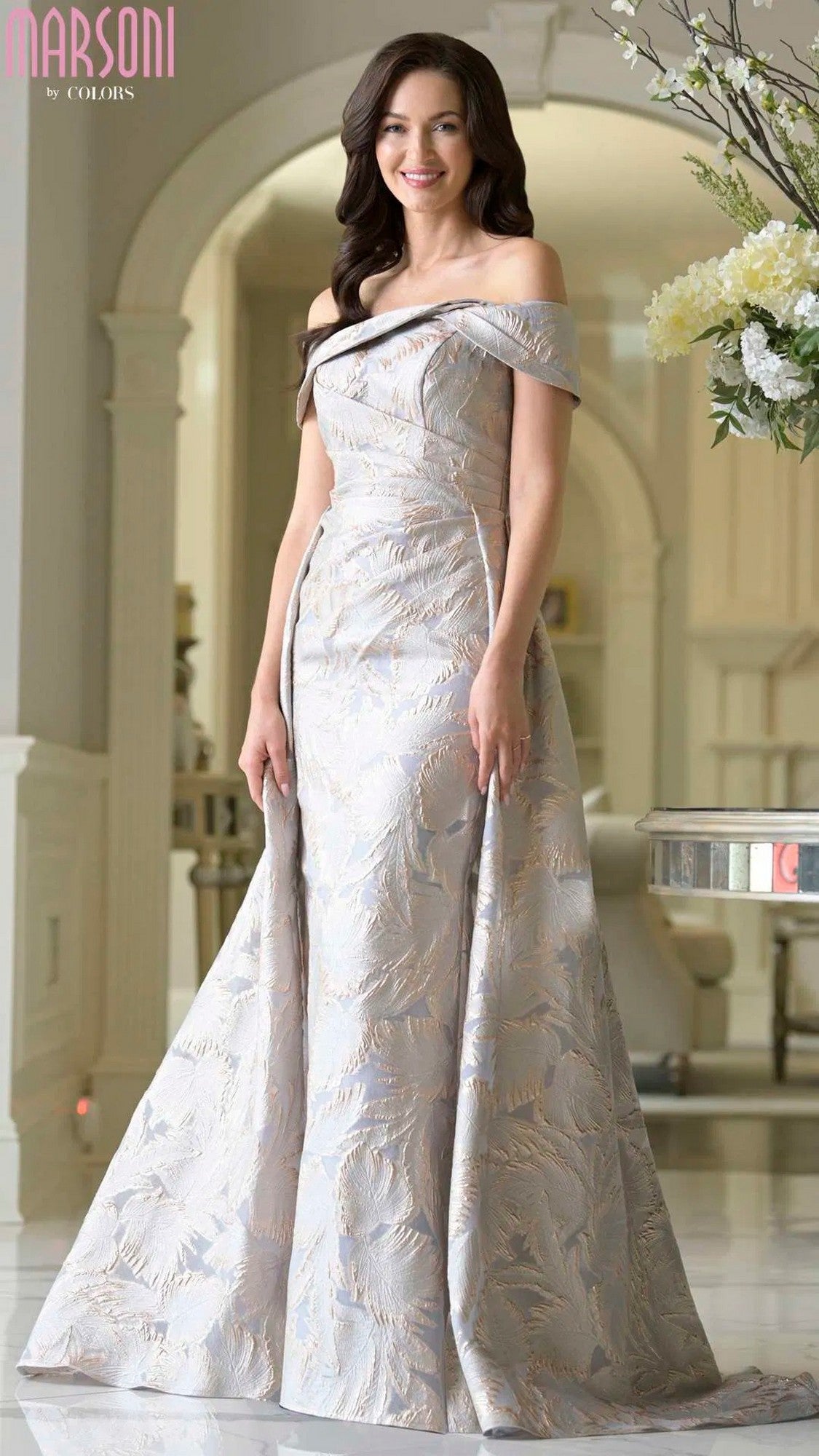 A woman in the Marsoni MV1382 long formal dress, featuring a gold gray patterned jacquard design, stands indoors and smiles at the camera.